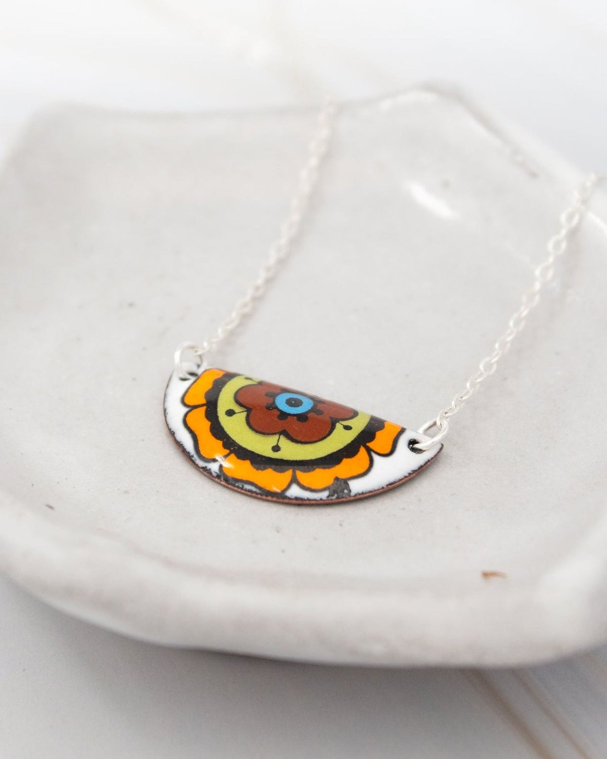 A colorful, half-circle pendant necklace with a floral design is displayed on a white plate.