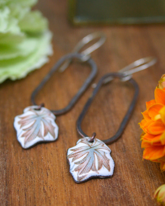 A pair of earrings with leaf-shaped pendants, one with a blue and white leaf design and the other with a brown leaf design, are displayed on a wooden surface alongside some flowers.
