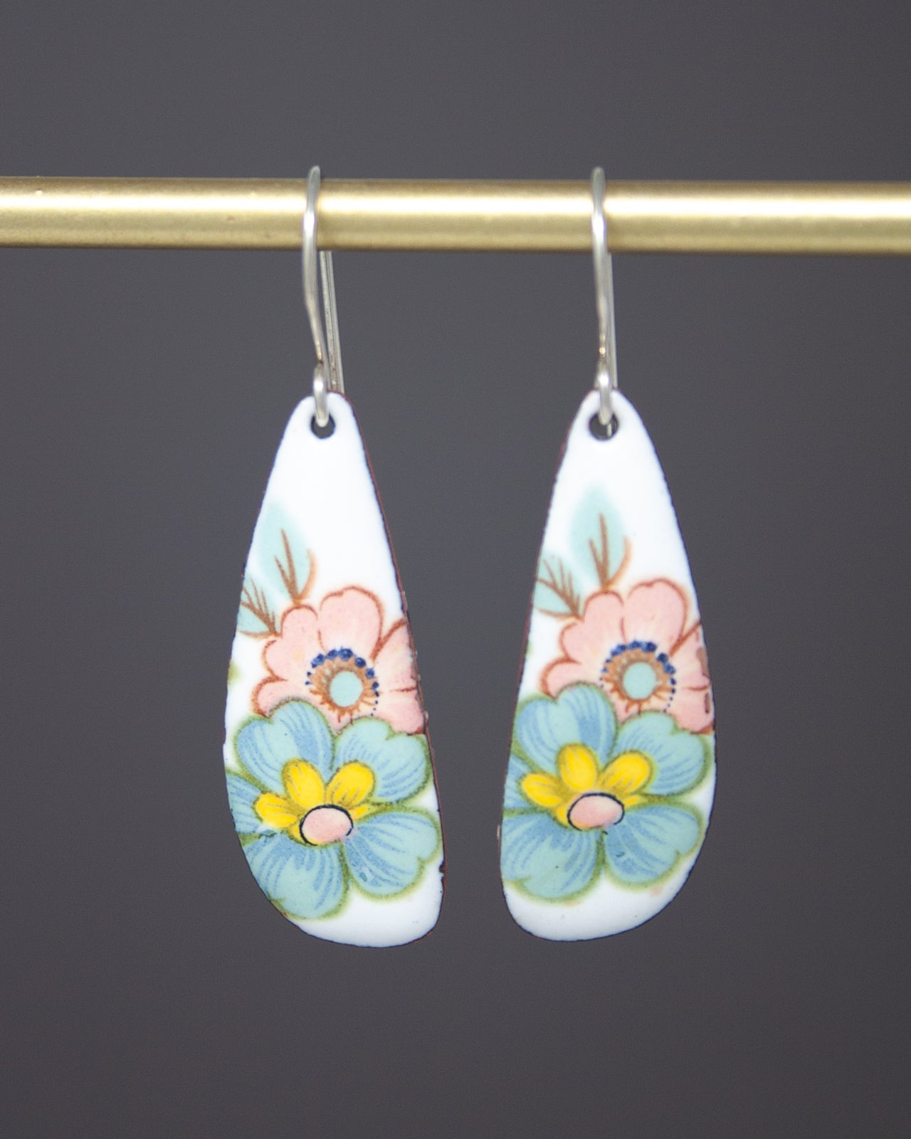 A pair of white earrings with colorful floral designs hanging from a gold bar.