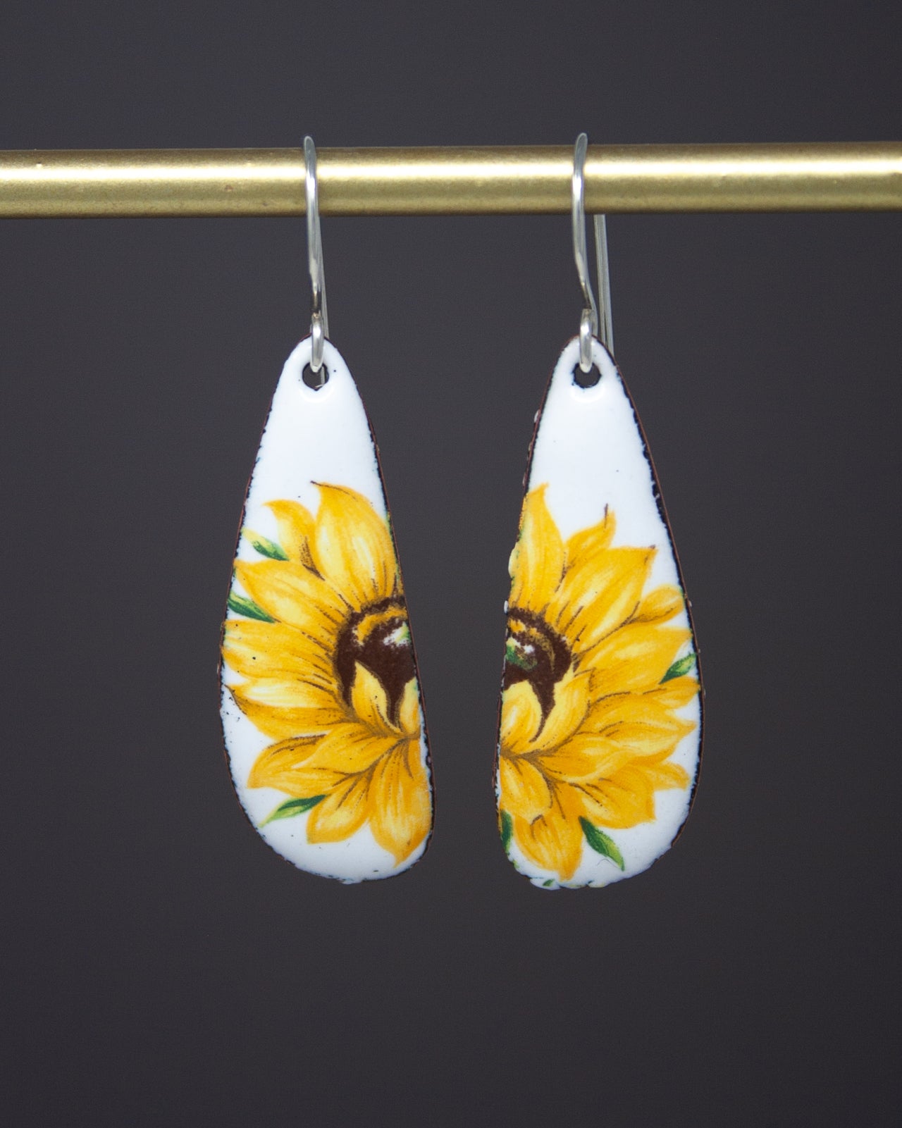 A pair of white earrings with yellow sunflower designs hanging from a gold bar.