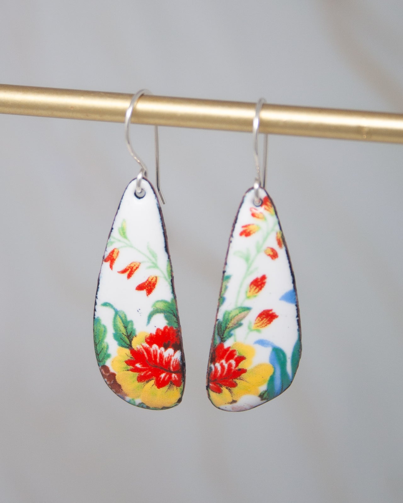 A pair of colorful earrings with floral designs hanging from a gold-colored metal bar.