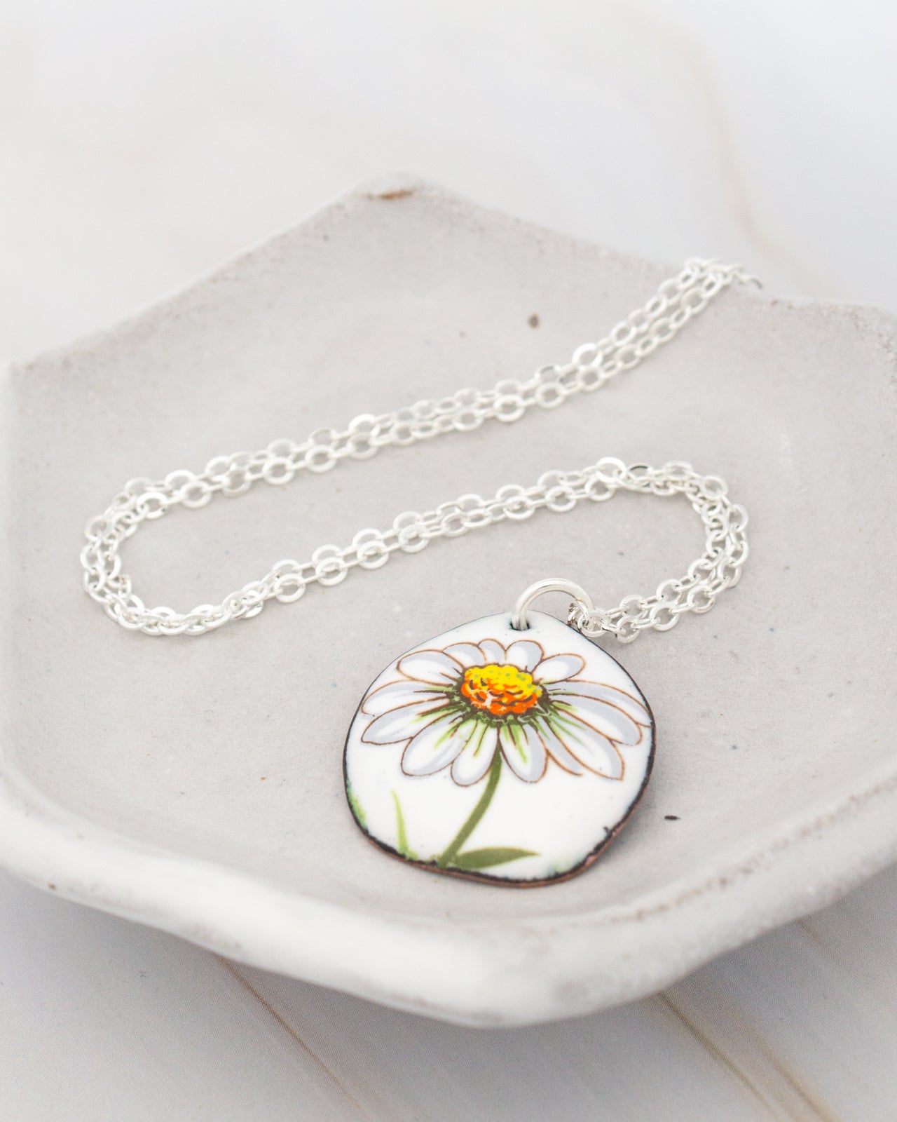 A necklace with a pendant featuring a white daisy flower design is displayed on a white plate.