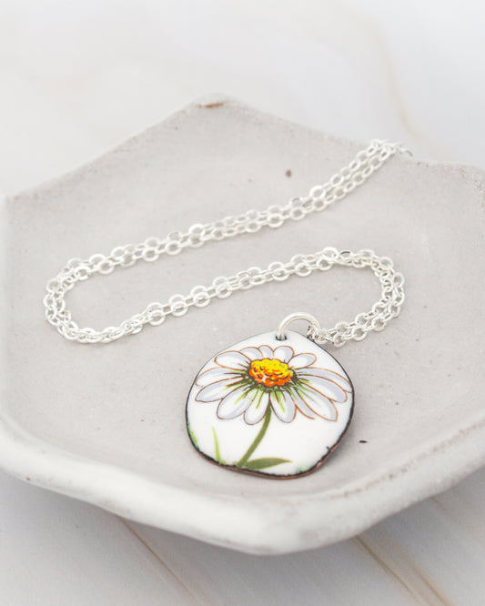 A necklace with a pendant featuring a white daisy flower design is displayed on a white plate.