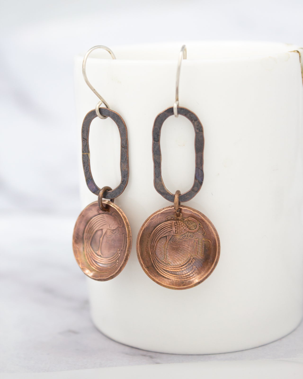 A pair of copper-colored earrings with circular pendants hanging from a white ceramic mug.