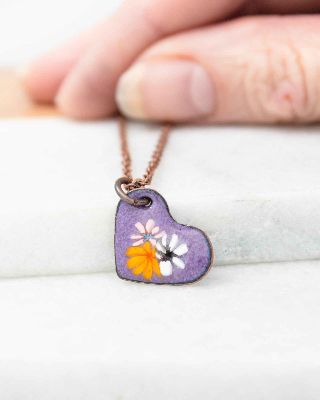 A hand is holding a small heart-shaped pendant necklace with a purple heart and orange and white flowers.