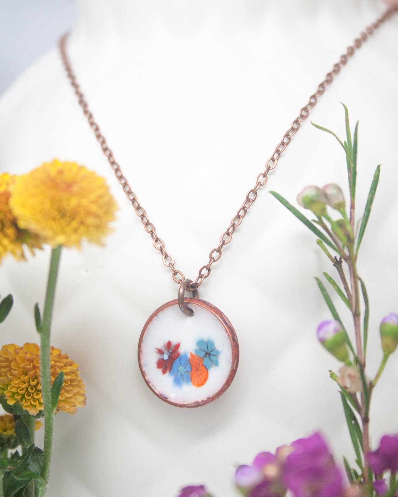 A necklace with a pendant featuring a floral design, surrounded by colorful flowers and a plant.