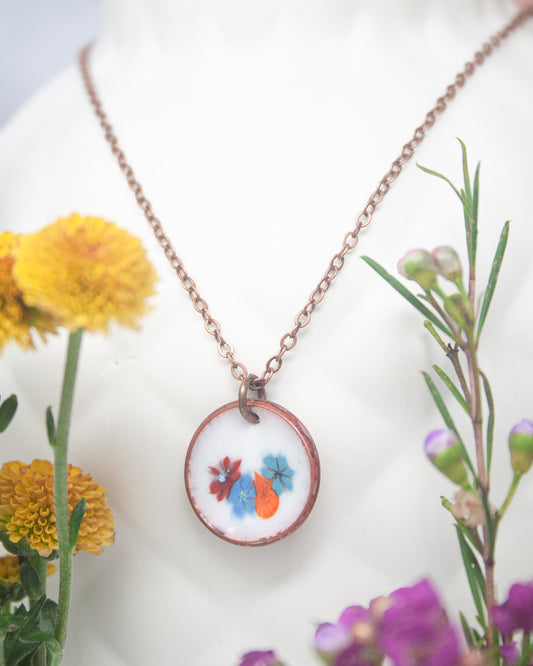 A necklace with a pendant featuring a floral design, surrounded by colorful flowers and a plant.