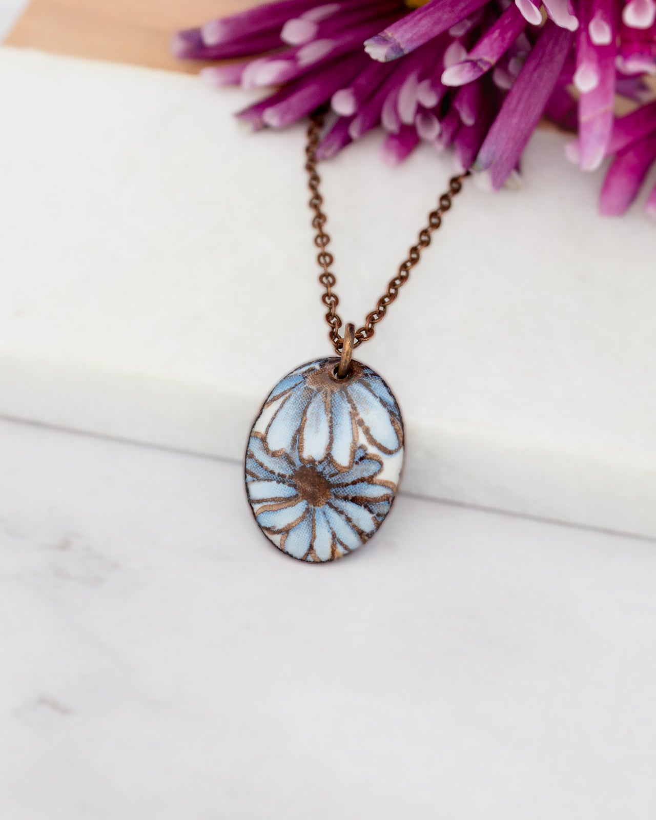 A necklace with a pendant featuring a blue and white flower design is displayed against a backdrop of purple flowers.