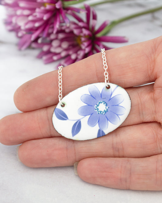 A hand holding a necklace with a blue flower design on it, with a blurred background of purple flowers.