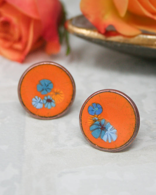 two round orange earrings with blue floral designs, placed on a white surface.
