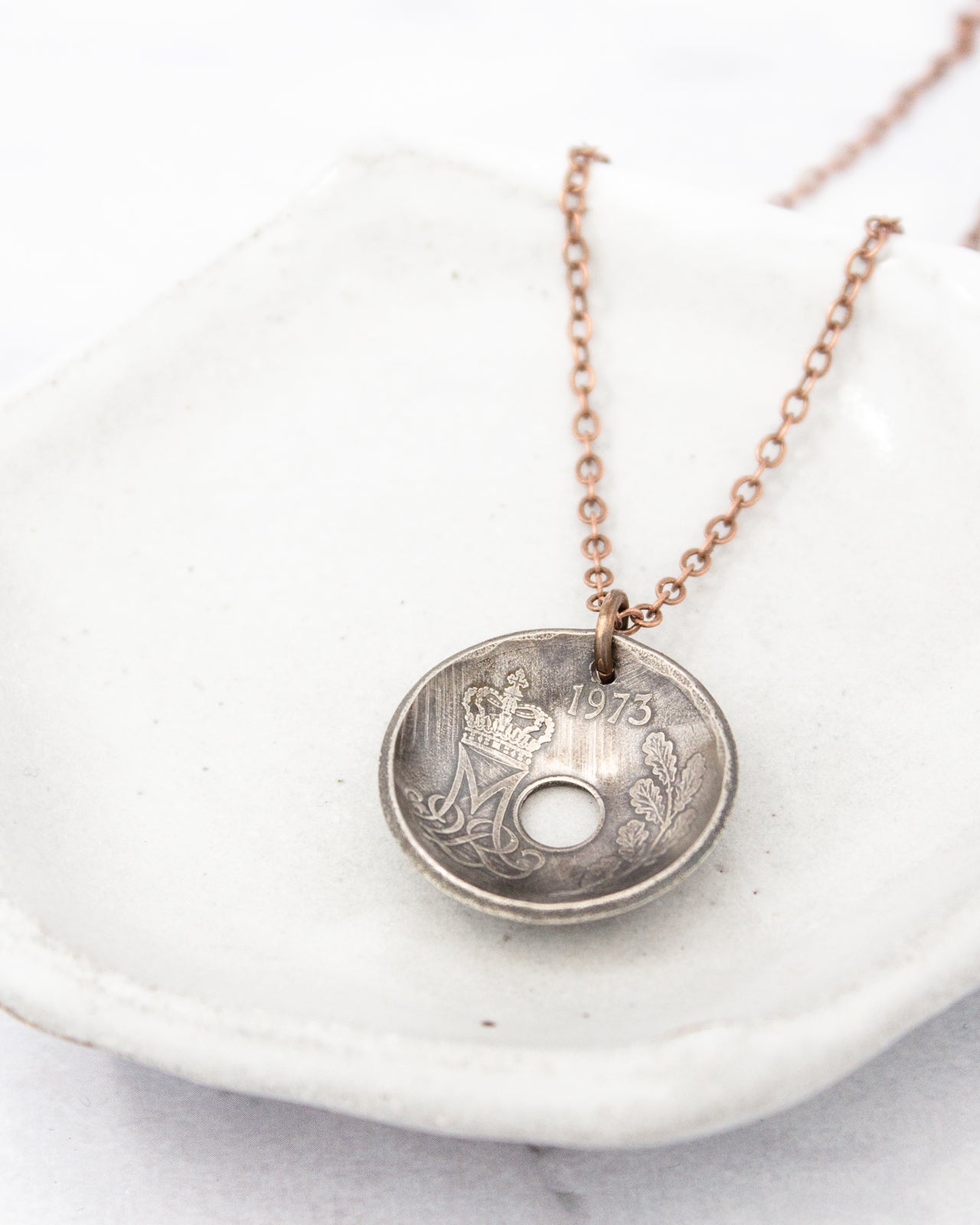 A necklace with a pendant made from a small, circular metal object, possibly a coin, with a design on it. The pendant is attached to a chain.