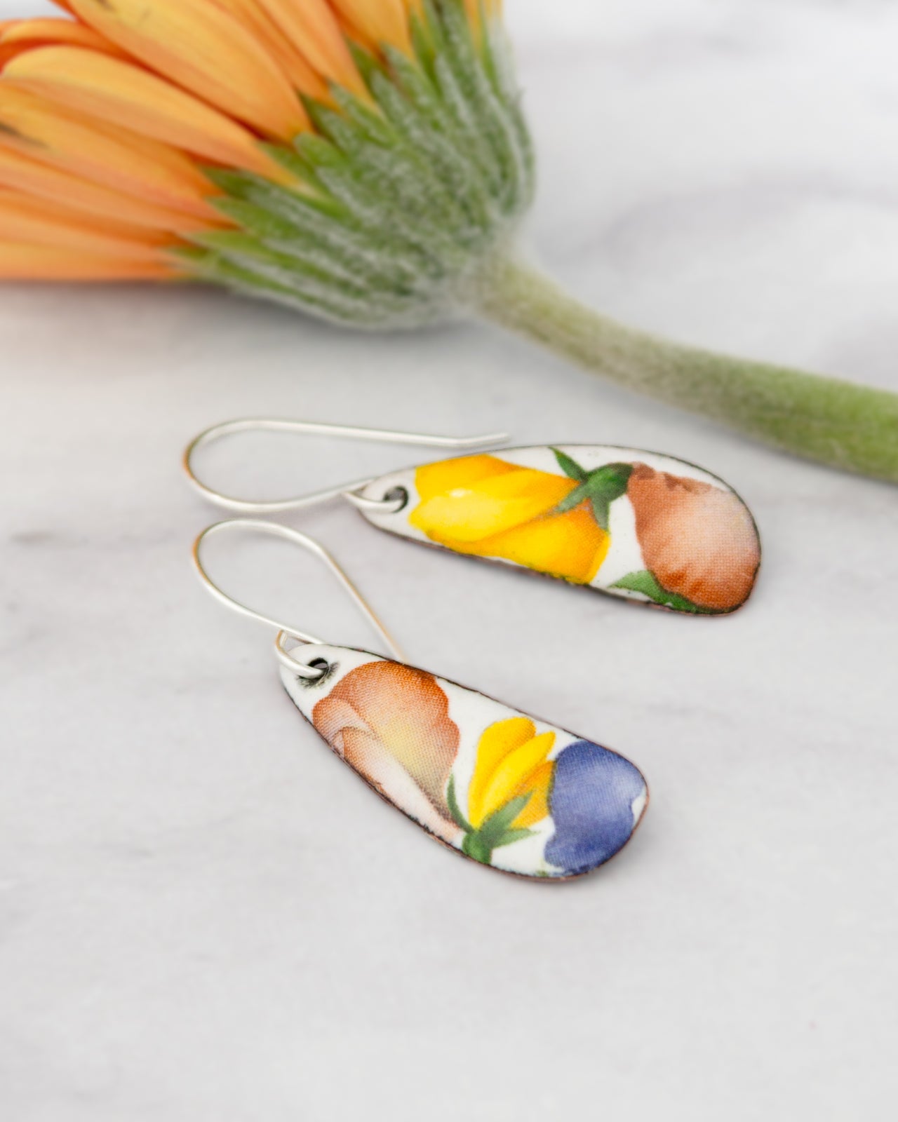 a pair of earrings with colorful floral designs, placed on a marble surface next to a flower.