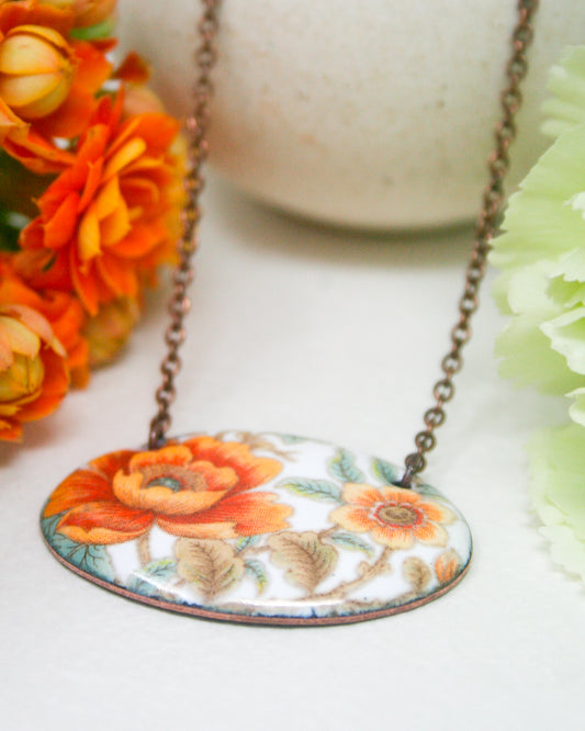 A necklace with a pendant featuring a floral design in shades of orange and white, resting on a white surface.