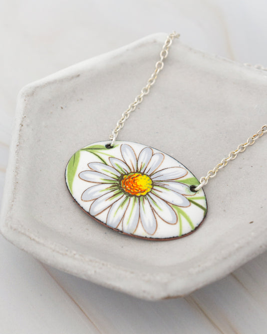 A necklace with a pendant featuring a white daisy flower with a yellow center, displayed on a white plate.