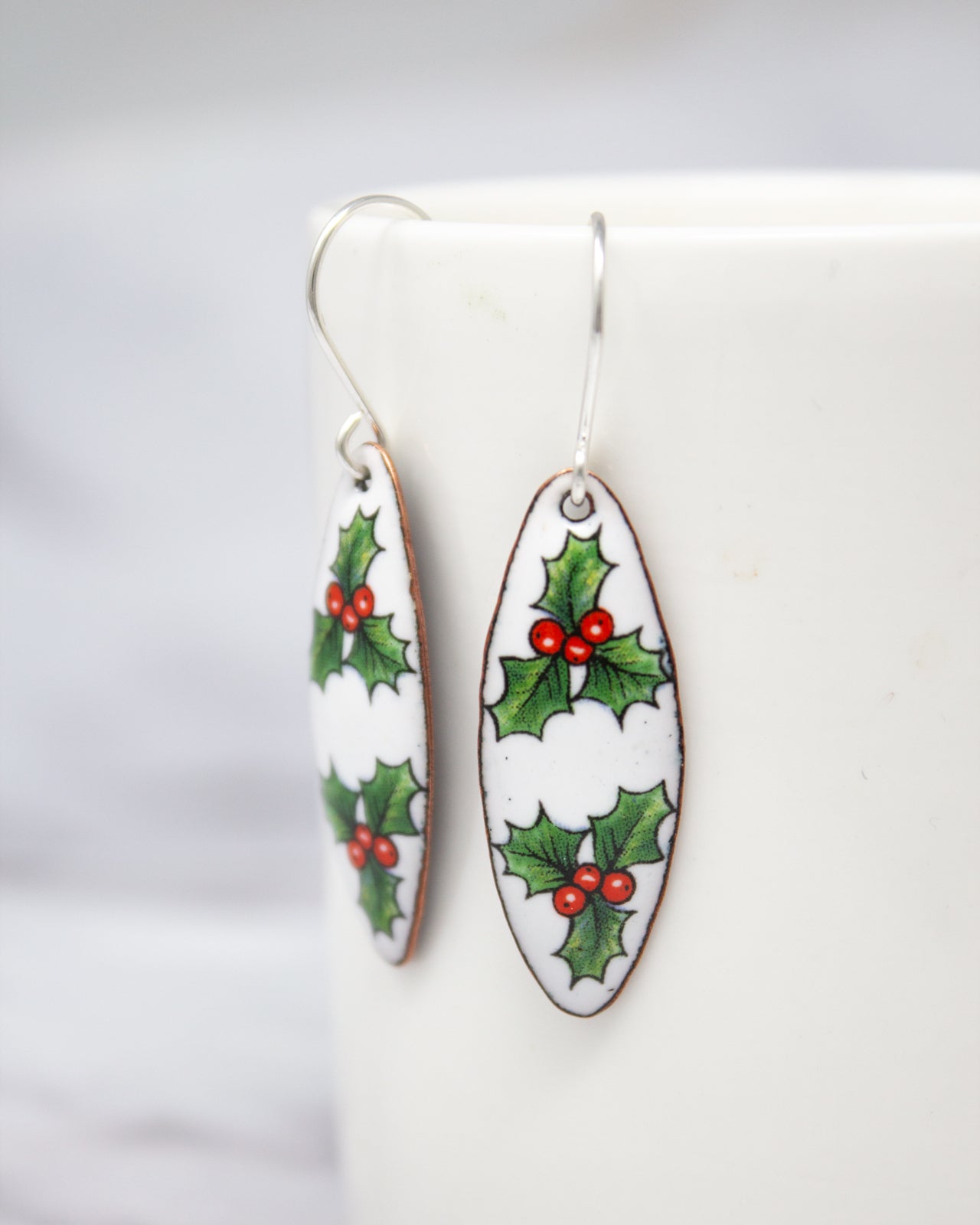 A pair of white earrings with red berries and green holly leaves on them, hanging from a hook.