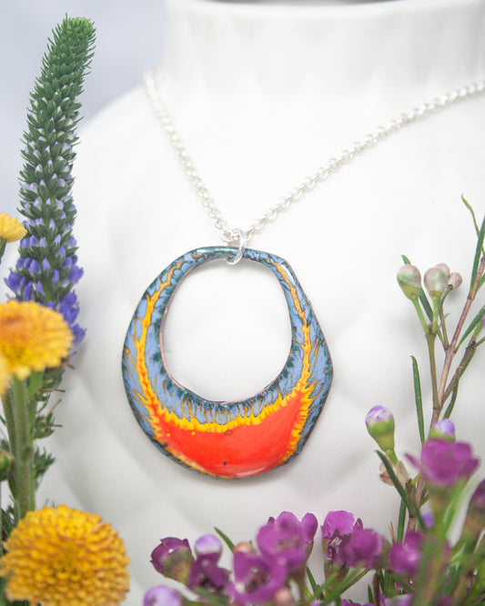 A colorful pendant necklace with a large, circular design hangs from a white bust form, surrounded by vibrant flowers and foliage.