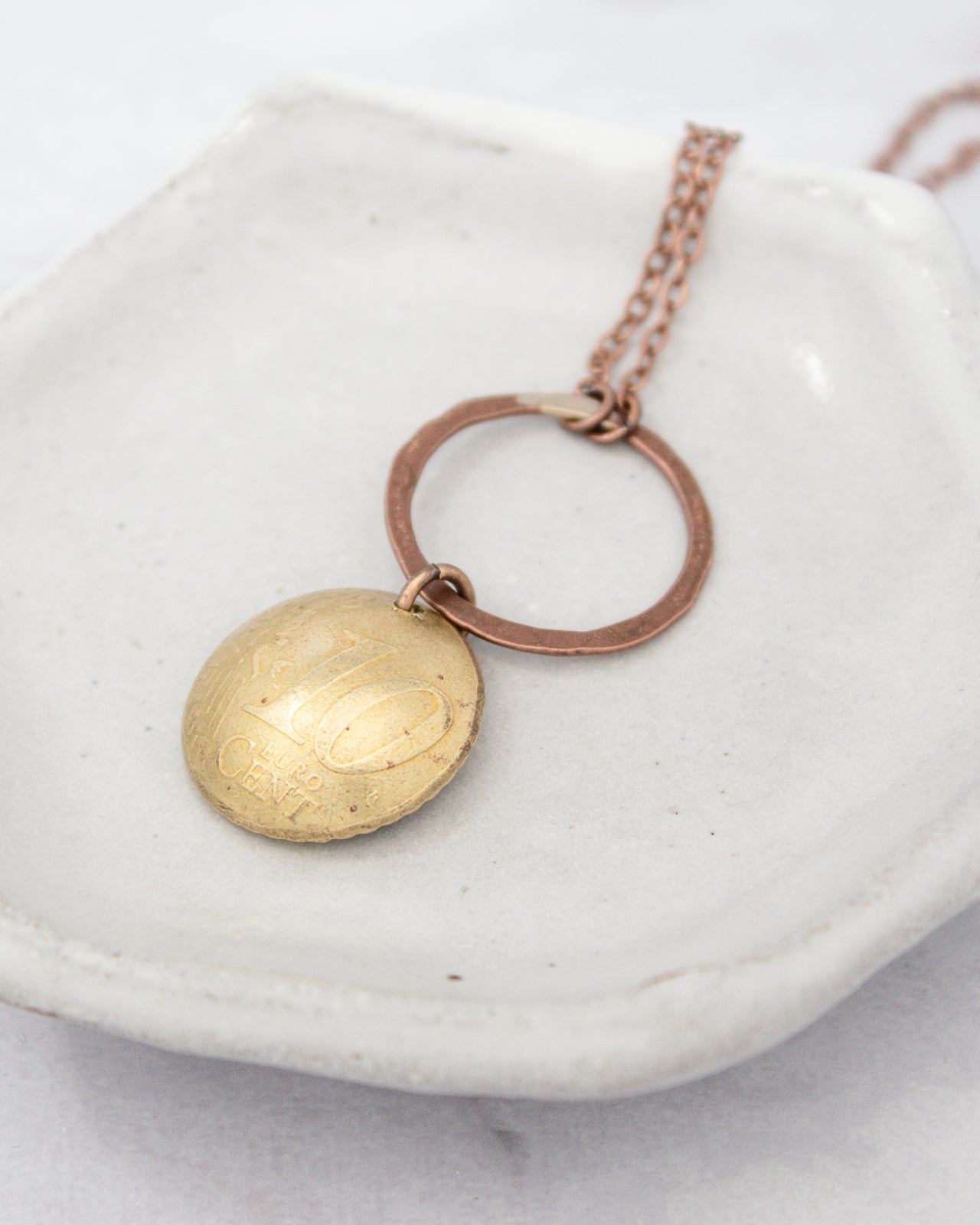 A gold-colored pendant with the inscription "10 CENTS" is hanging from a chain, resting on a white ceramic dish.