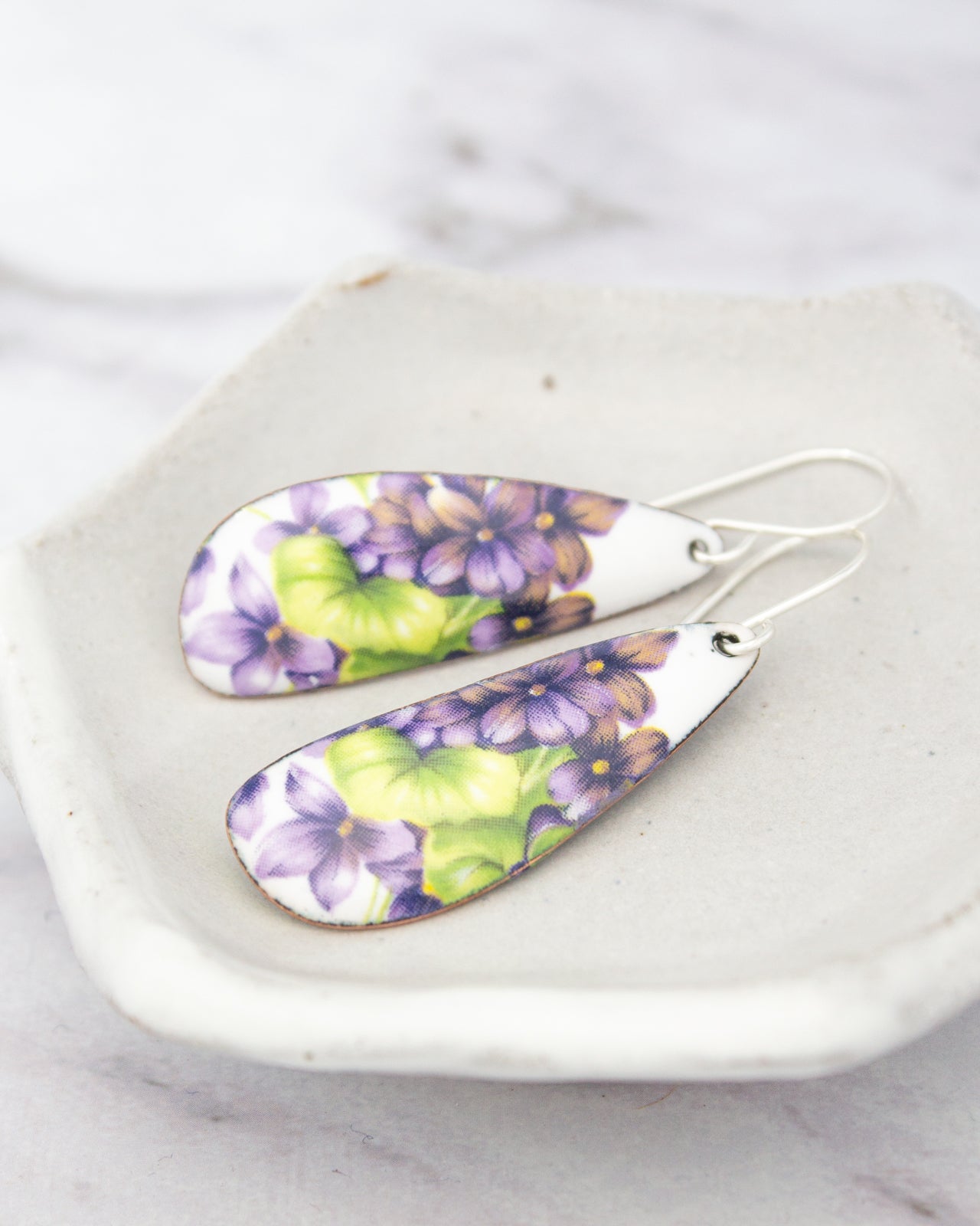 A pair of earrings with a floral design, featuring purple and green flowers, are displayed on a white plate.