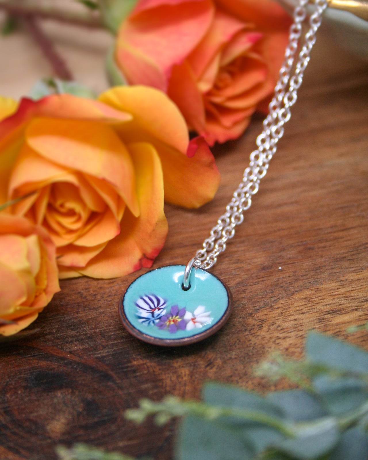 A necklace with a blue pendant featuring a floral design rests on a wooden surface, surrounded by vibrant orange and yellow roses.