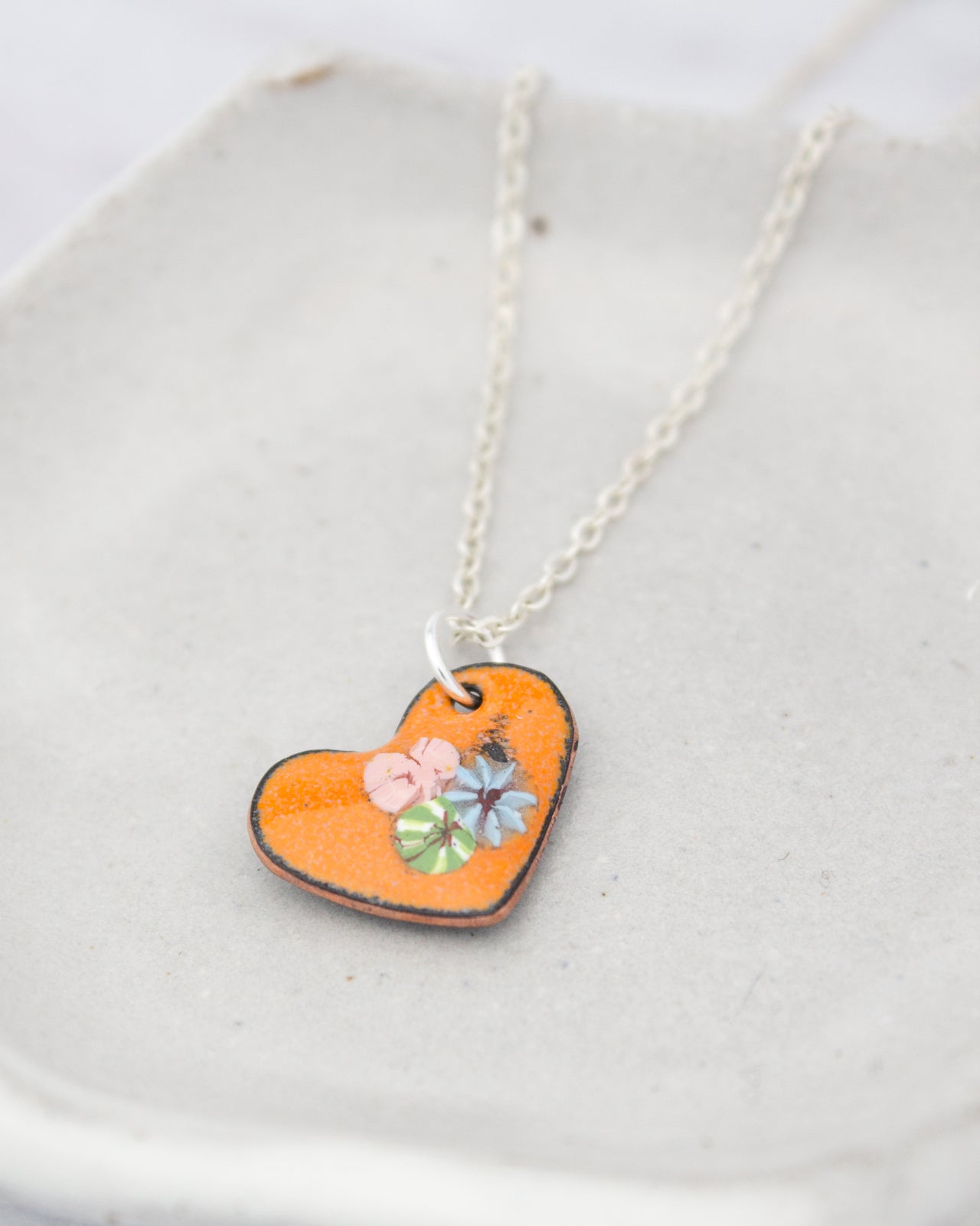 A heart-shaped pendant with a floral design is displayed on a white surface, attached to a silver chain.