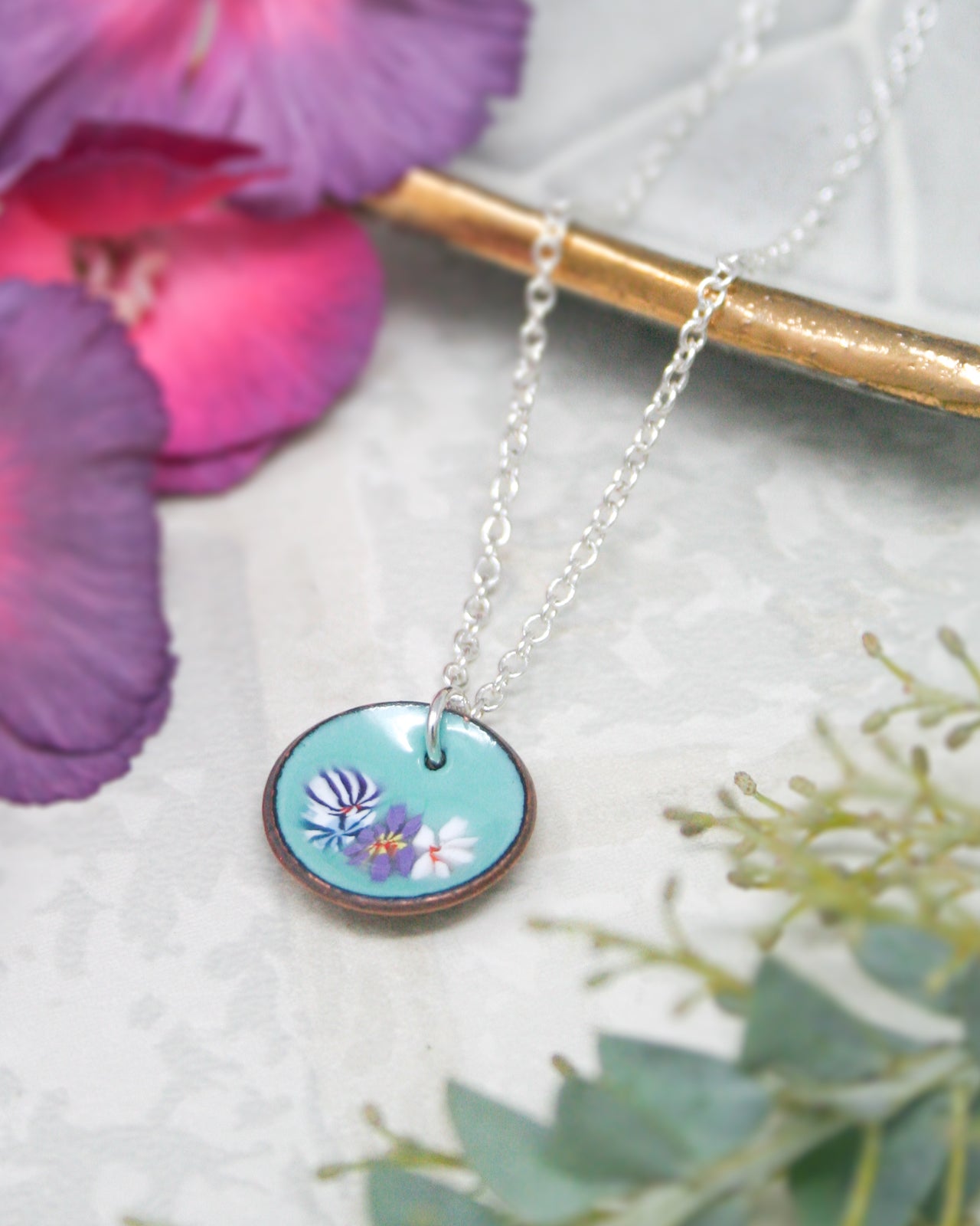 A necklace with a circular pendant featuring a floral design is displayed on a white surface, surrounded by purple and green flowers.