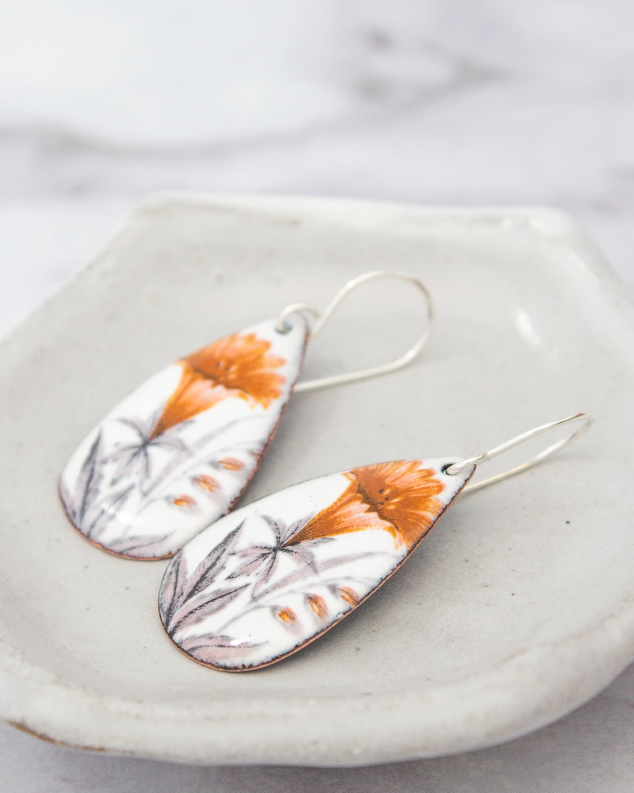 A pair of earrings with a floral design, featuring orange and white flowers, resting on a white plate.