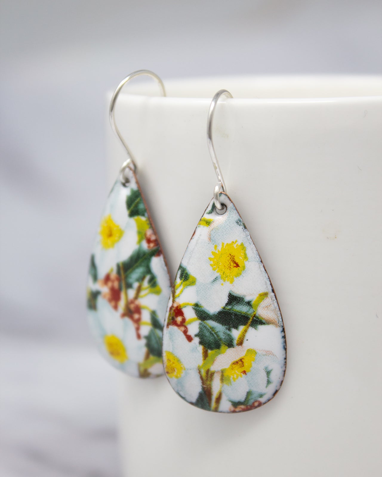 A pair of earrings with a floral pattern, featuring yellow flowers and green leaves, are displayed on a white surface.