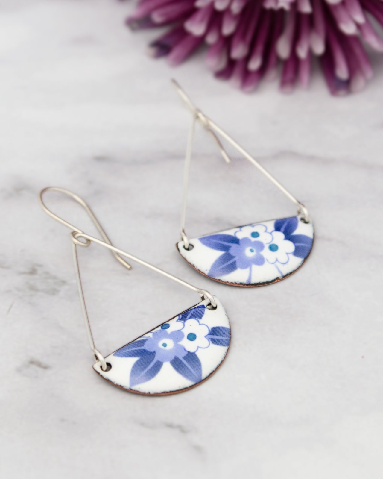 A pair of silver earrings with blue floral designs, placed on a marble surface next to a purple flower.