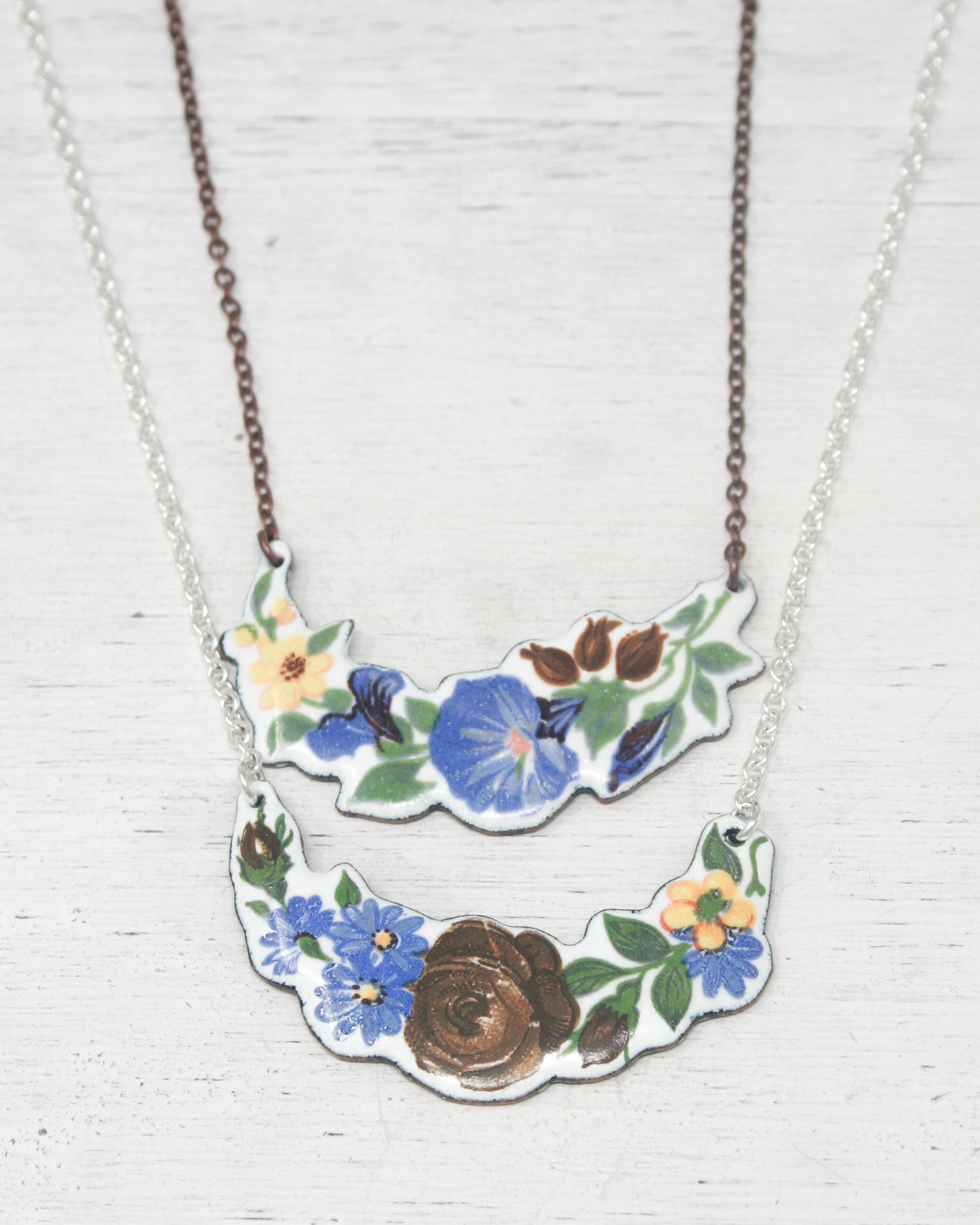 two necklaces with floral designs. The necklaces have a combination of blue, yellow, and brown colors, and they are displayed on a white wooden surface.