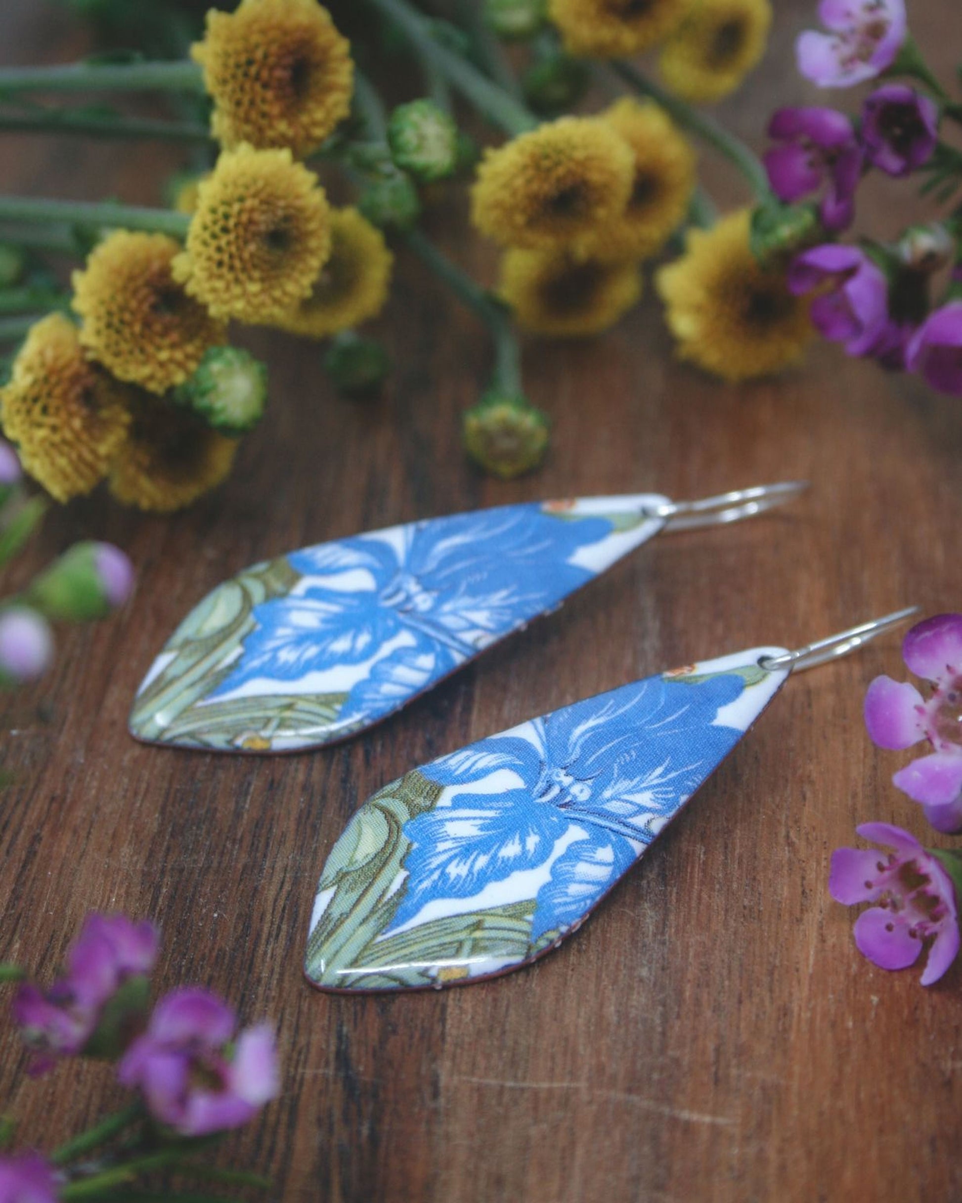 A pair of blue and white floral earrings rests on a wooden surface, surrounded by yellow flowers and purple flowers.