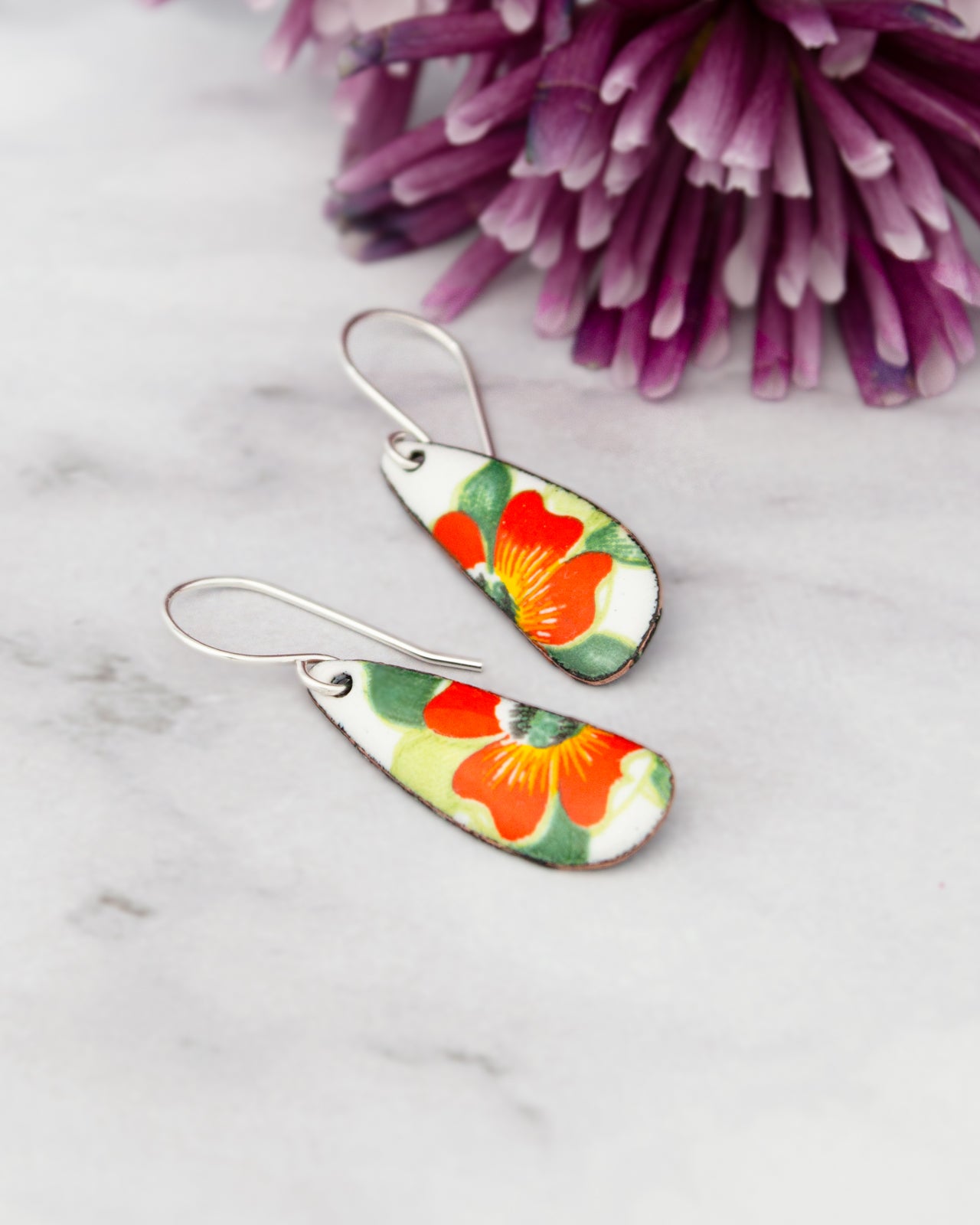 A pair of earrings with a floral design, featuring red and orange flowers, are placed on a white marble surface next to a purple flower.
