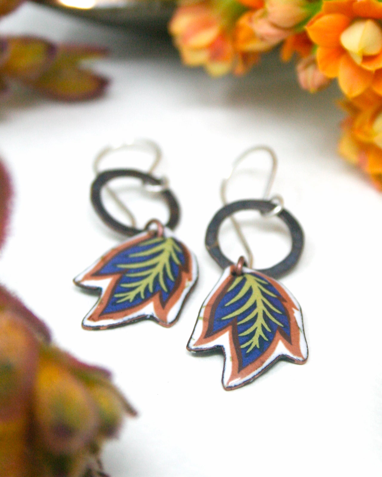 A pair of colorful earrings with leaf-shaped designs, placed on a white surface with orange flowers in the background.