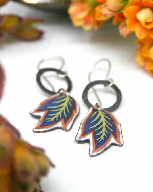 A pair of colorful earrings with leaf-shaped designs, placed on a white surface with orange flowers in the background.