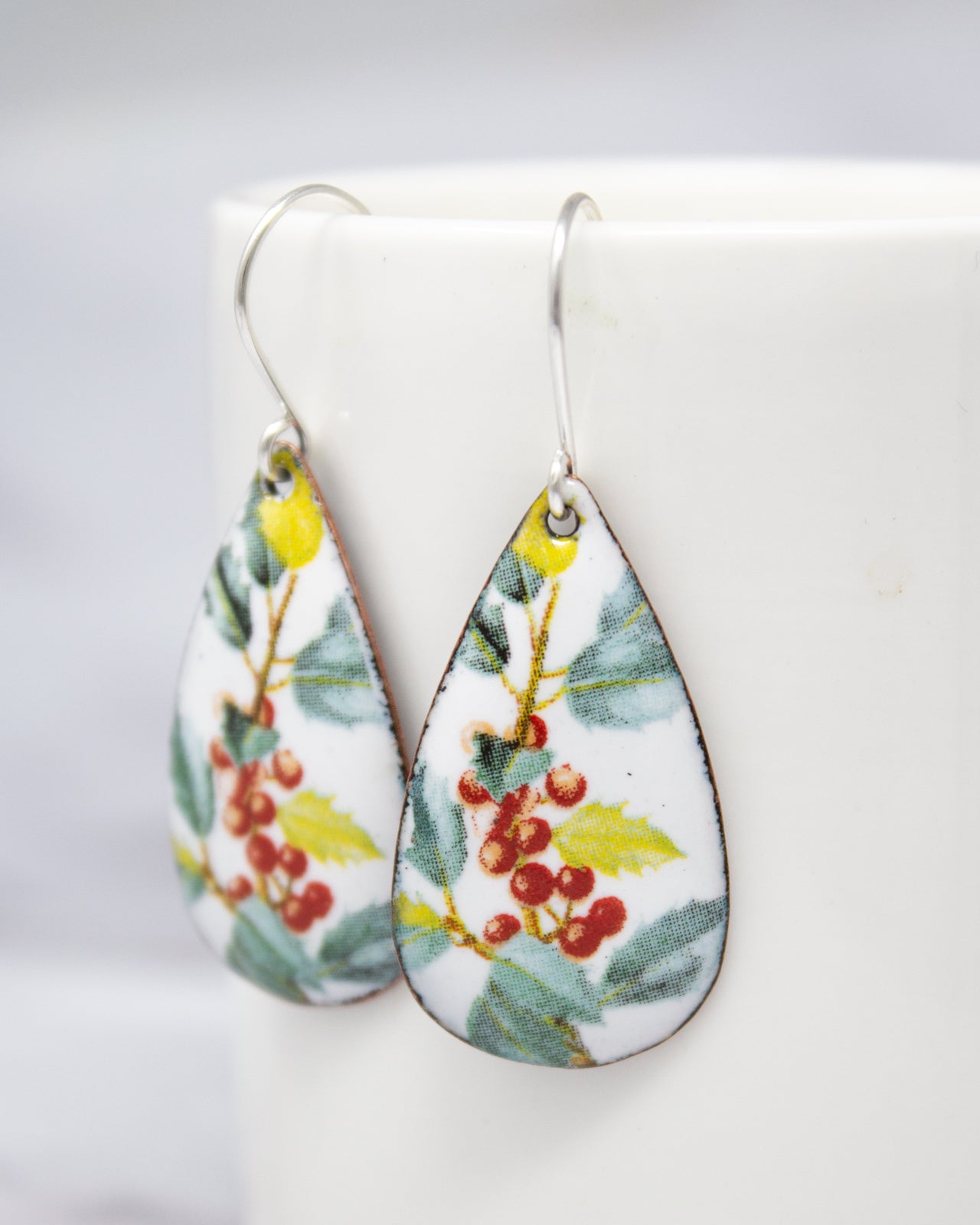 A pair of earrings with a floral design, featuring red berries and green leaves, are displayed on a white surface.