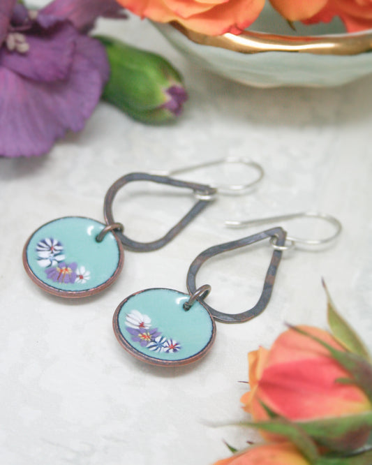 A pair of turquoise earrings with floral designs, placed on a white surface alongside colorful flowers and a bowl.