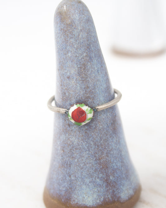 A ring with a red flower design is displayed on a blue ceramic stand.