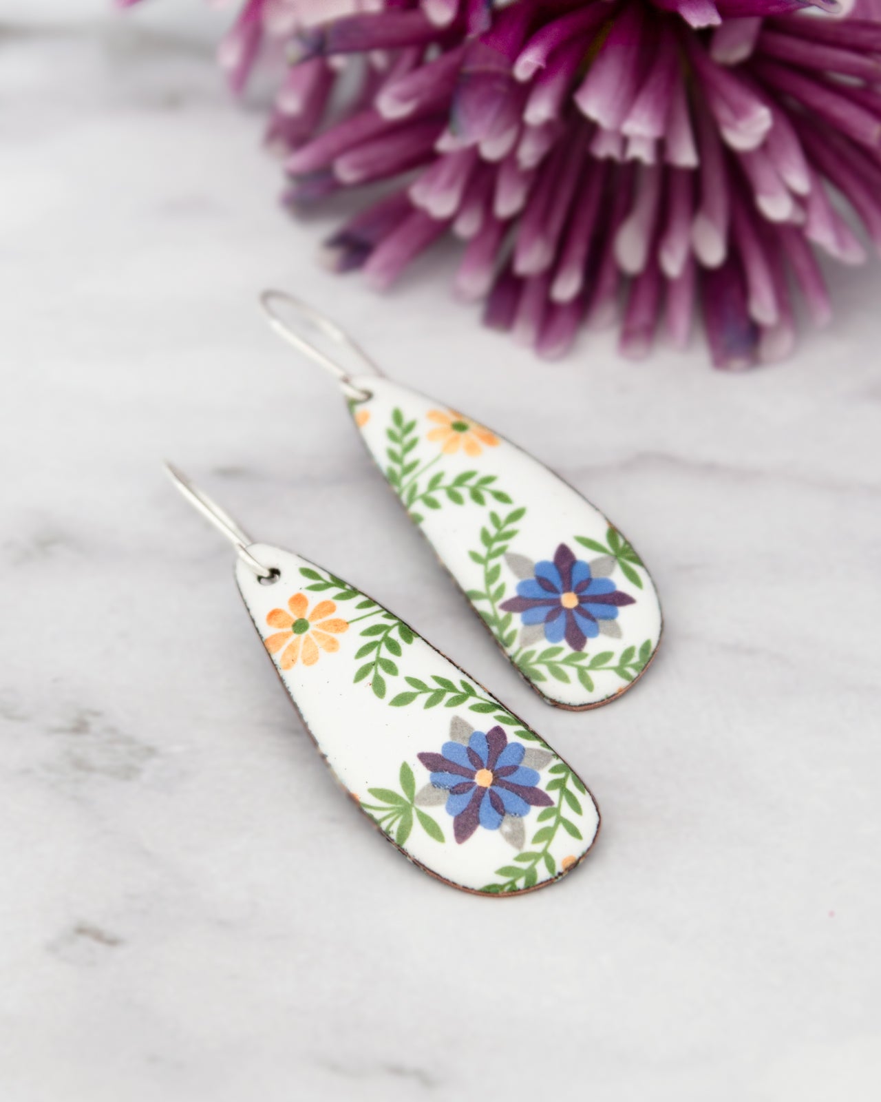 two white earrings with colorful floral designs, placed on a marble surface next to a vibrant purple flower.