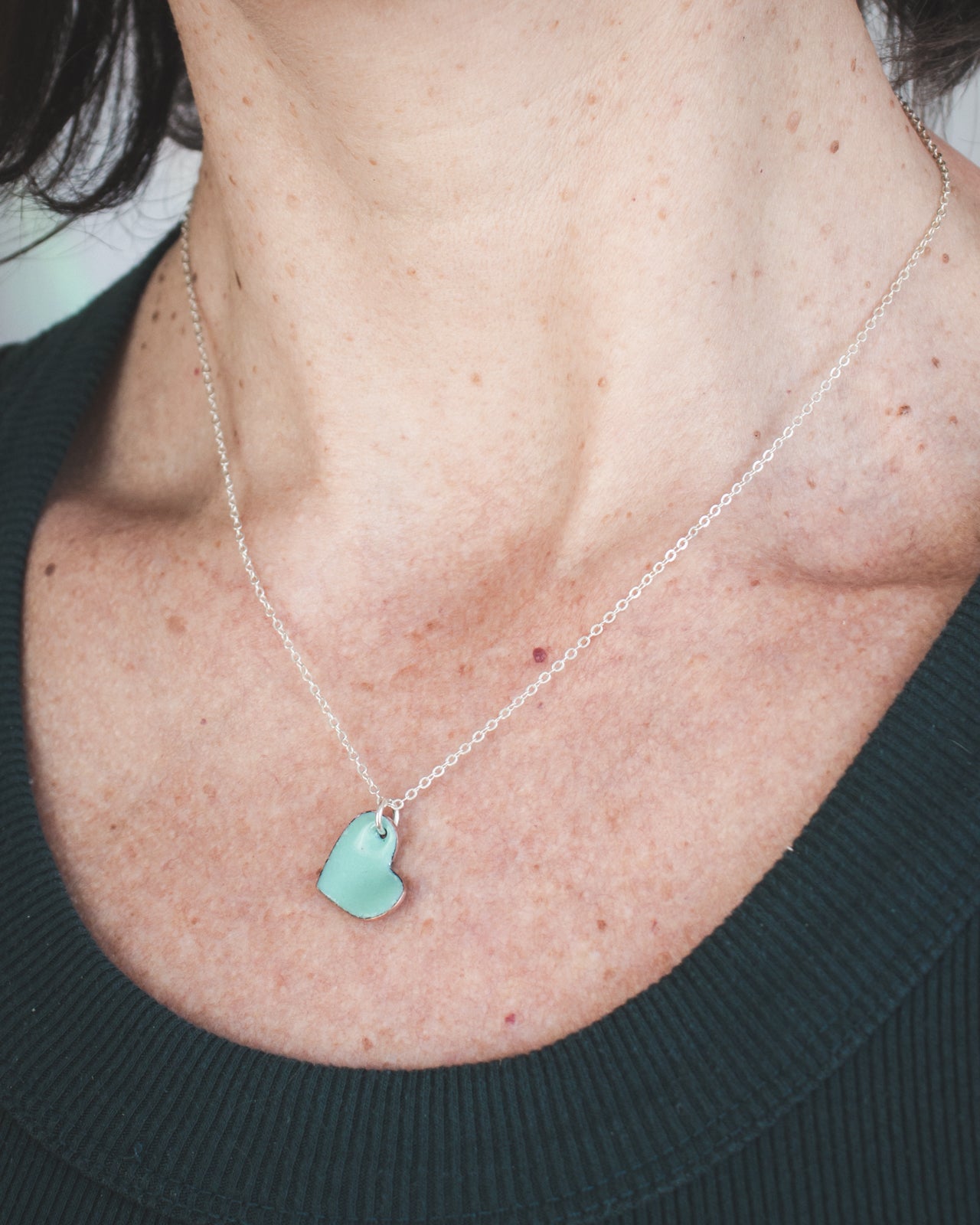 A woman wearing a silver necklace with a small turquoise heart-shaped charm.