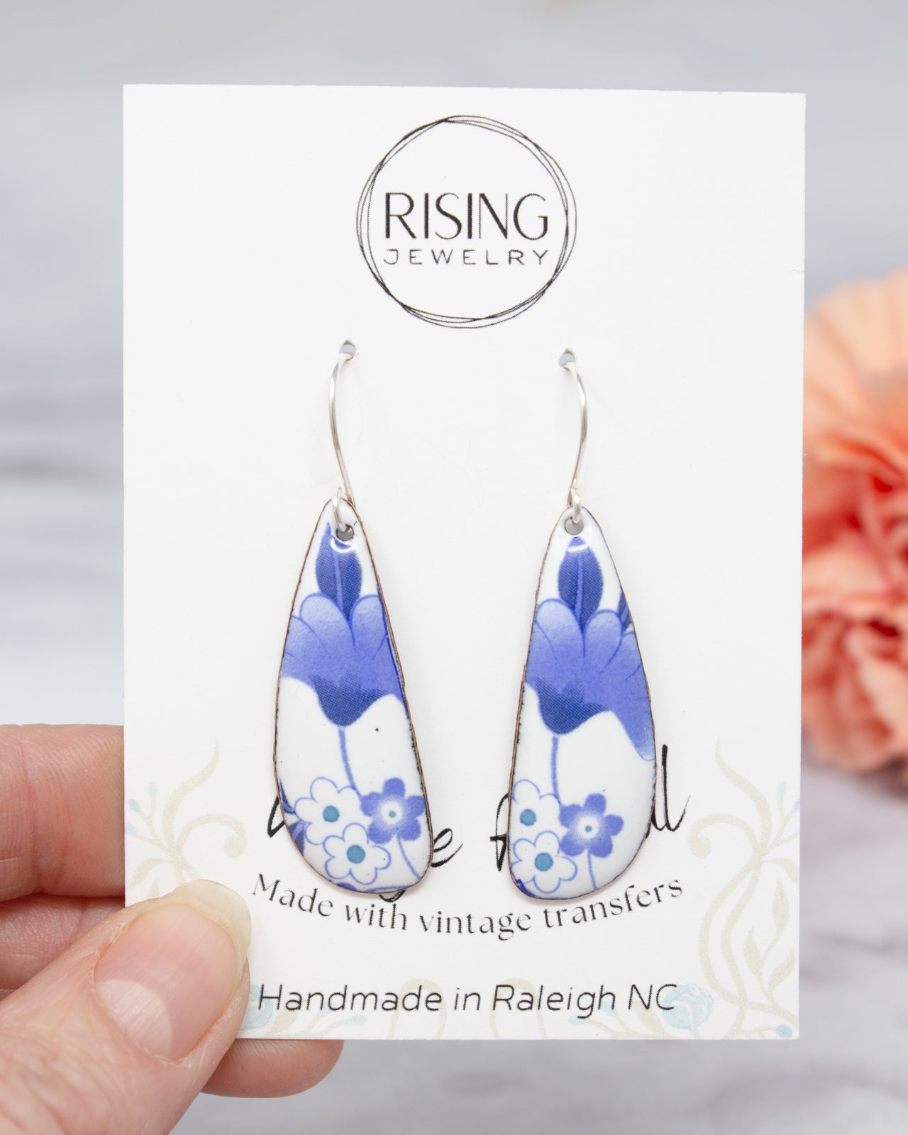 A hand holding a pair of earrings with a floral design, against a blurred background.