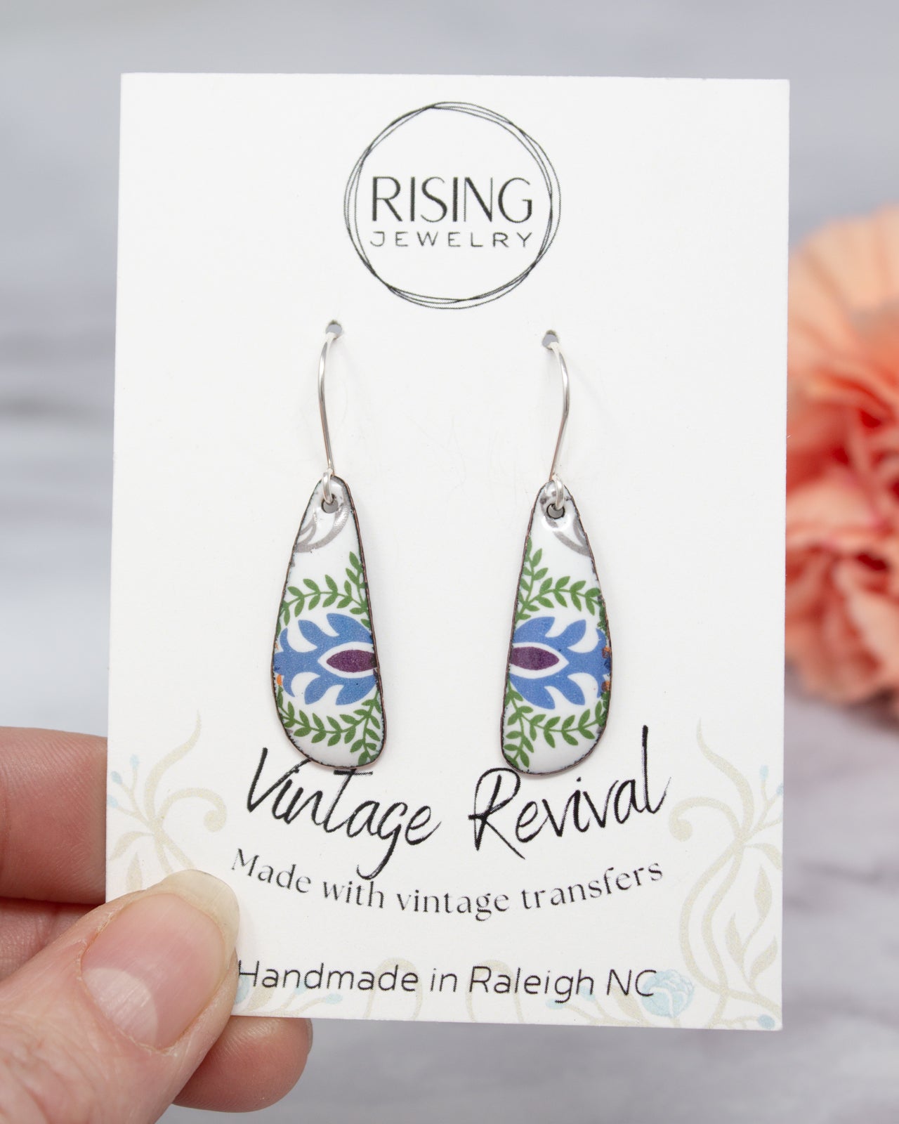 A hand holding a pair of earrings with a card that displays the earrings and the company's name.