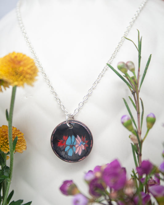 A necklace with a circular pendant featuring a floral design is displayed against a backdrop of yellow and purple flowers.