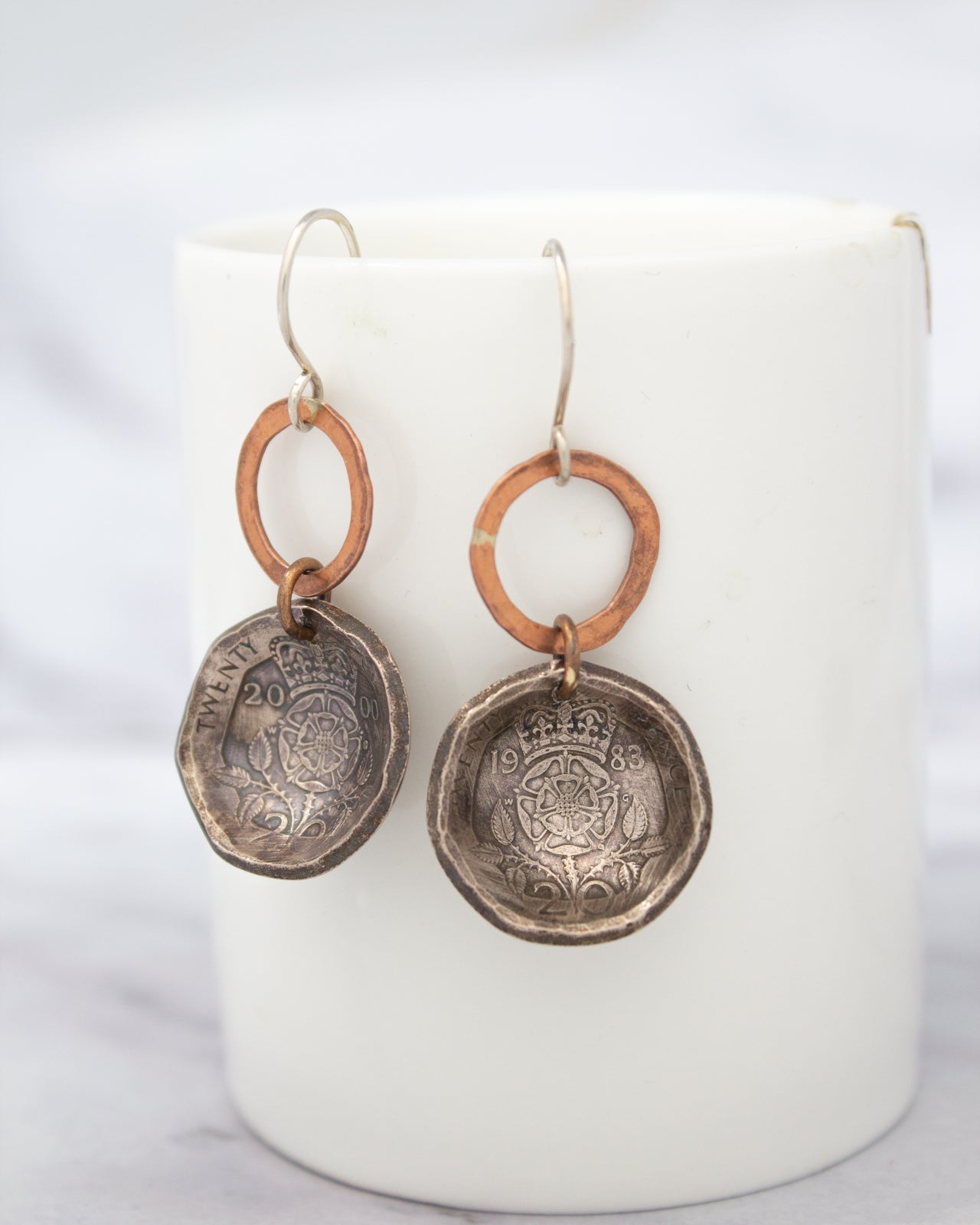 A pair of earrings made from a silver-colored metal, featuring a circular design with a floral pattern and a small metal charm hanging from it.