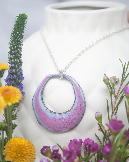 A necklace with a large, colorful pendant is displayed in a white vase, surrounded by a variety of colorful flowers.