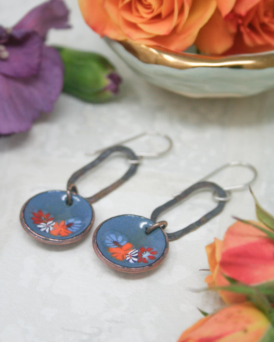 a pair of blue earrings with floral designs, placed on a white surface next to a bowl of orange roses.