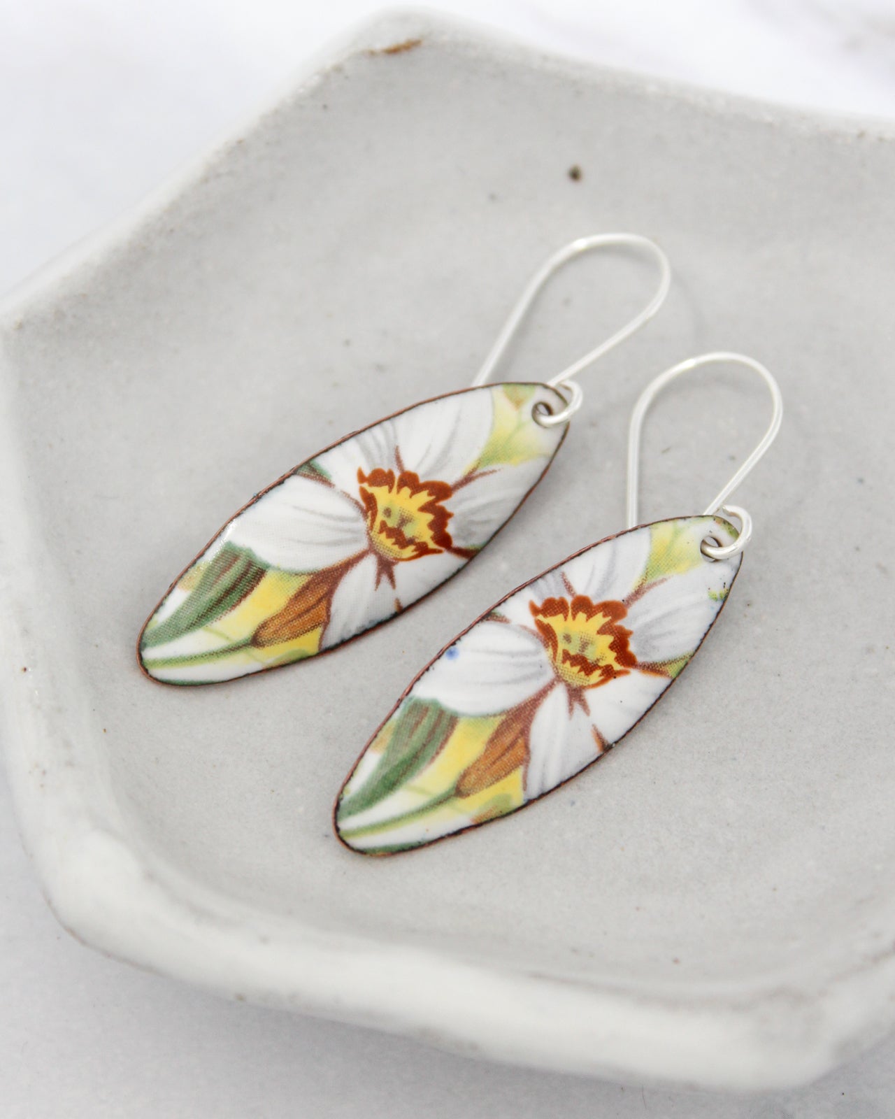 A pair of earrings with a floral design, featuring white and yellow flowers, resting on a white surface.