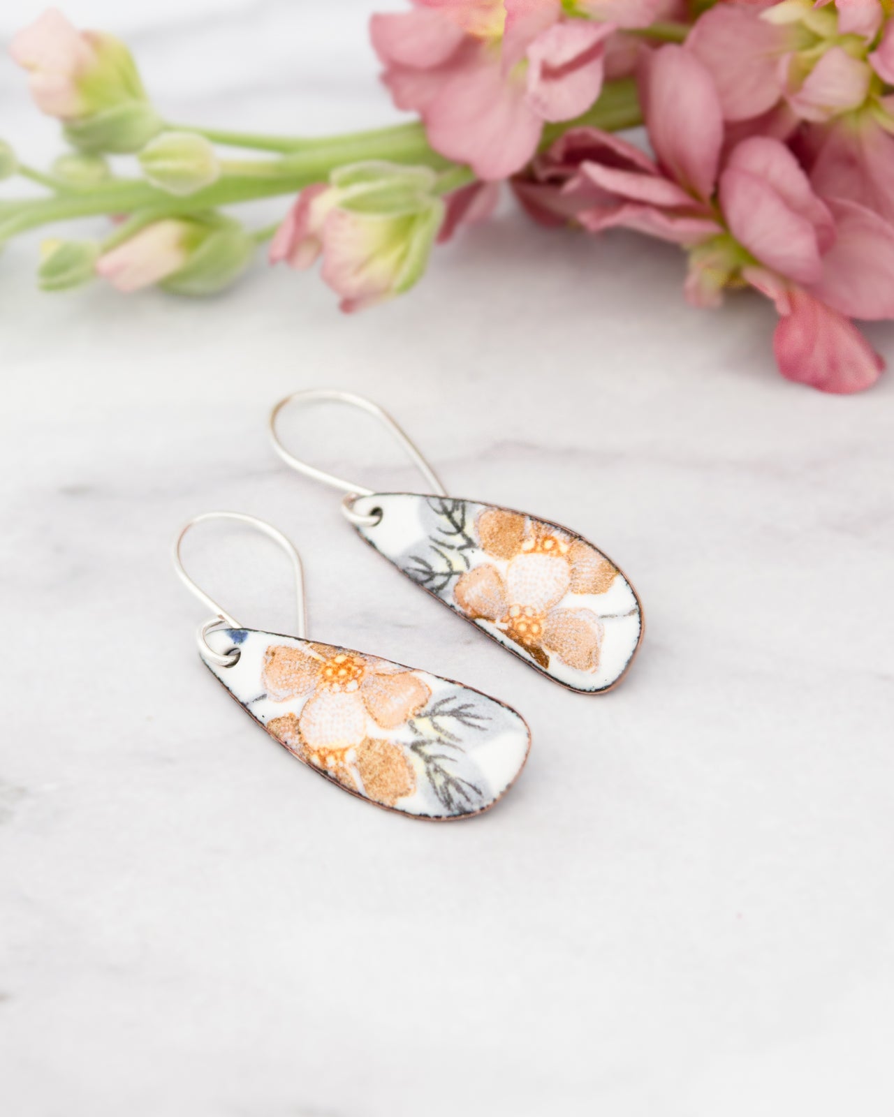 A pair of earrings with a floral design, placed on a marble surface next to a bouquet of pink flowers.