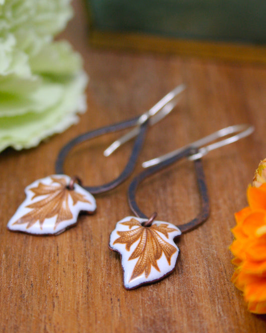 a pair of earrings with leaf-shaped pendants, placed on a wooden surface alongside some flowers.