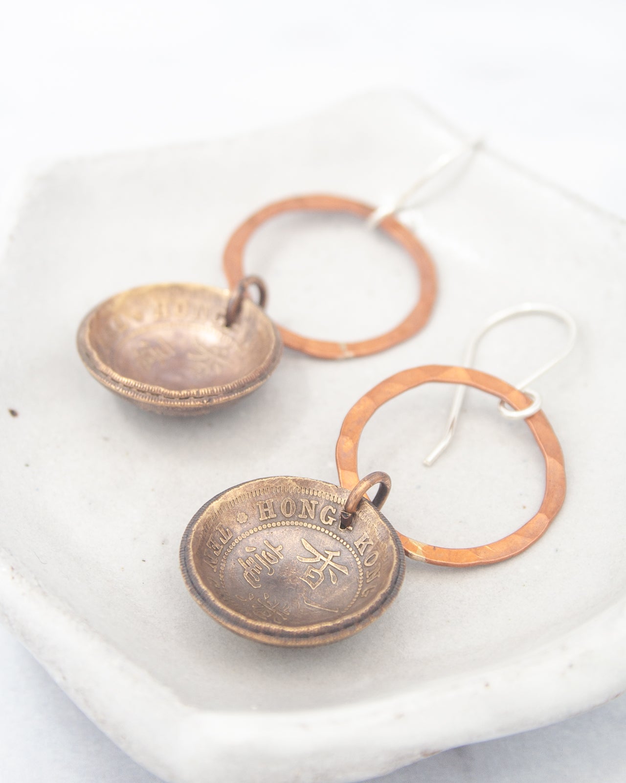 A pair of earrings made of two metal pieces, one with a circular design and the other with a ring-like shape, resting on a white surface.