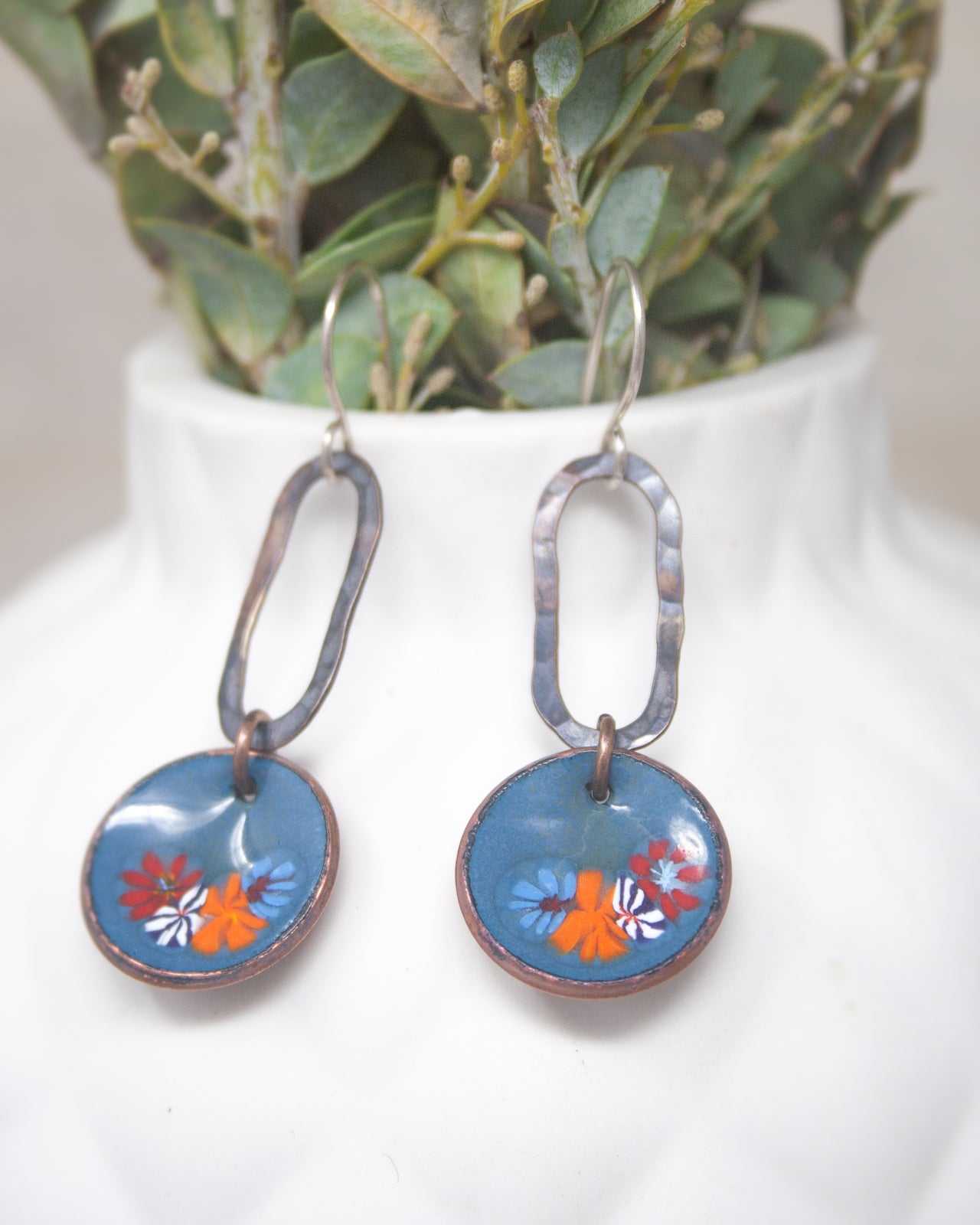 A pair of blue earrings with a floral design hangs from a hook, displayed in front of a white vase with greenery.