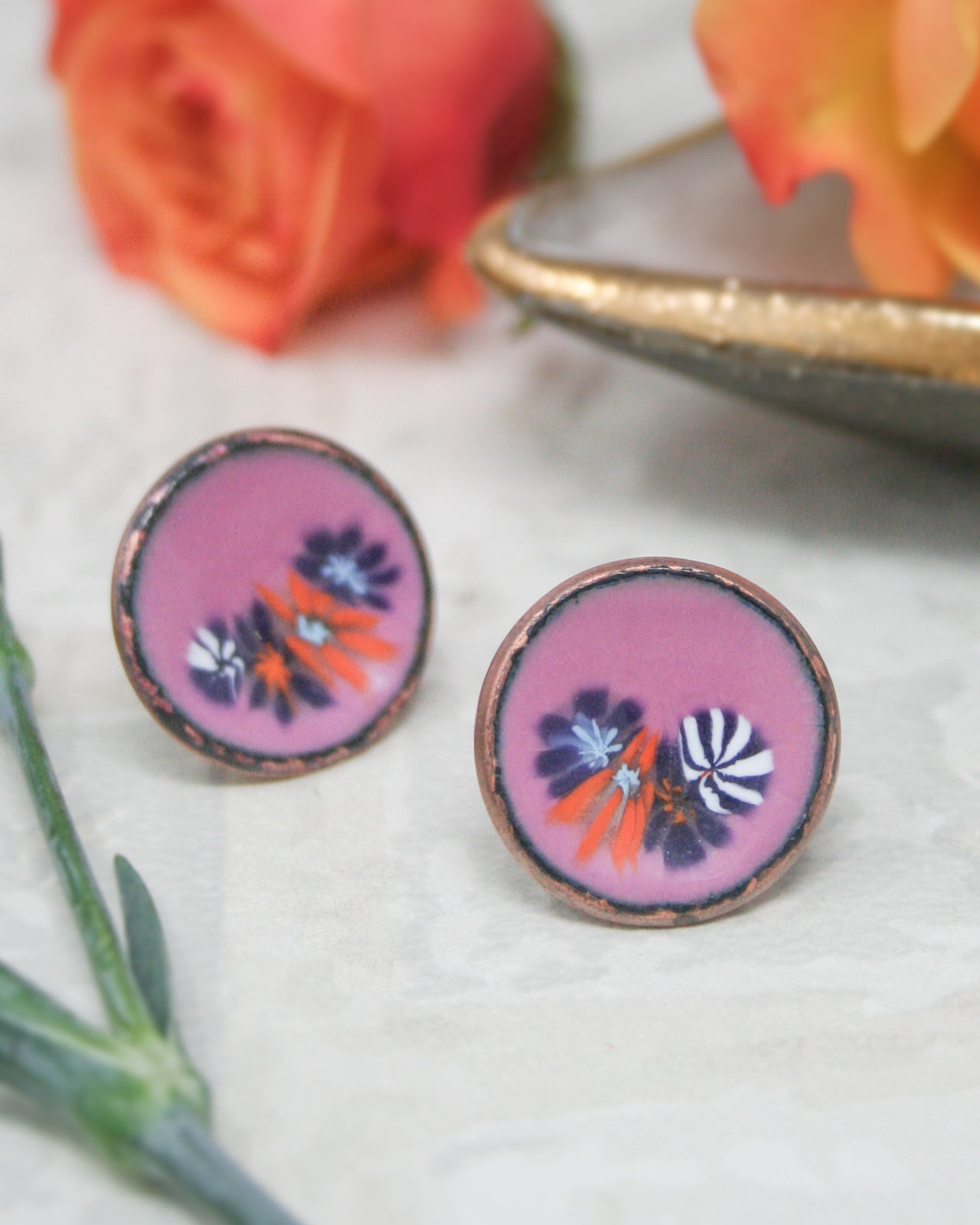 two round earrings with a floral design, placed on a white surface alongside a pink rose and a green leaf.